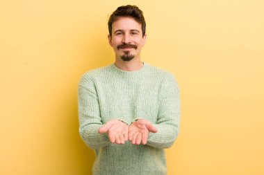 Genç İspanyol adam mutlu bir şekilde gülümsüyor, arkadaş canlısı, kendinden emin, olumlu bakışlı, bir nesne ya da konsept sunuyor ve gösteriyor.