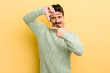 Genç İspanyol adam mutlu, arkadaş canlısı ve pozitif hissediyor, gülümsüyor ve elleriyle bir portre veya fotoğraf çerçevesi yapıyor.