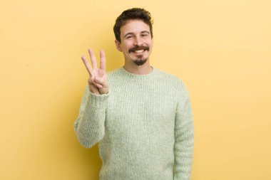 Genç İspanyol adam gülümsüyor ve arkadaş canlısı görünüyor, üç ya da üç numarayı gösteriyor, eli önde, geriye doğru sayıyor.