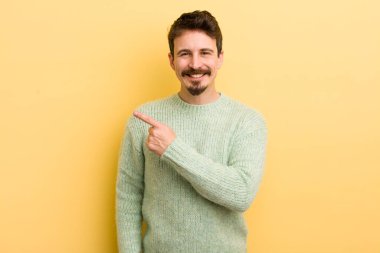 Genç İspanyol adam neşeyle gülümsüyor, mutlu hissediyor ve kenarı ve yukarıyı işaret ediyor, fotokopi alanında nesneler gösteriyor.