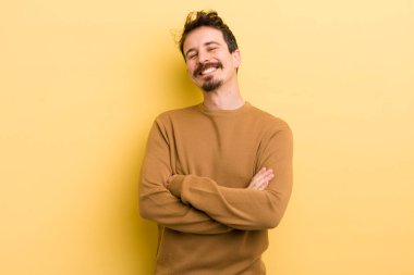 Genç İspanyol adam kollarını kavuşturup mutlu bir şekilde gülüyor, rahat, pozitif ve tatmin edici bir pozla.