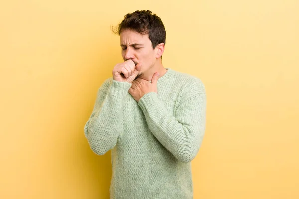 stock image young hispanic man feeling ill with a sore throat and flu symptoms, coughing with mouth covered