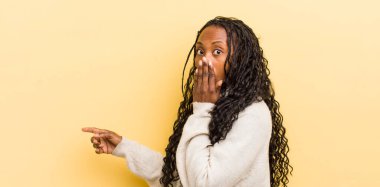 Siyah, güzel bir kadın mutlu, şok olmuş ve şaşırmış hissediyor. Ağzını eliyle kapatıyor ve yanal fotokopi alanını işaret ediyor.