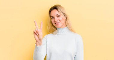 Sarışın, beyaz bir kadın gülümsüyor ve arkadaş canlısı görünüyor. İki ya da iki numarayı gösteriyor. Elleri önde, geri sayıyor.