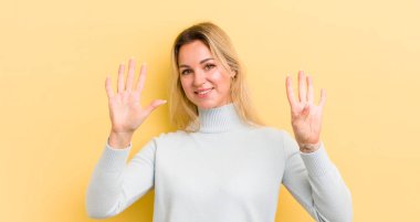 Sarışın, beyaz bir kadın gülümsüyor ve dost canlısı görünüyor. 9 ya da 9 numarayı gösteriyor. Eli önde, geri sayıyor.