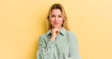 Sarışın, beyaz bir kadın mutlu görünüyor ve gülümsüyor, çenesi açık, merak ediyor ya da soru soruyor, seçenekleri karşılaştırıyor.