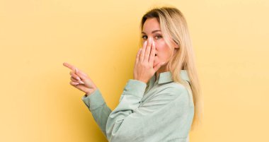 Sarışın, beyaz kadın kendini mutlu, şok olmuş ve şaşırmış hissediyor. Ağzını eliyle kapatıyor ve yanal fotokopi alanını işaret ediyor.
