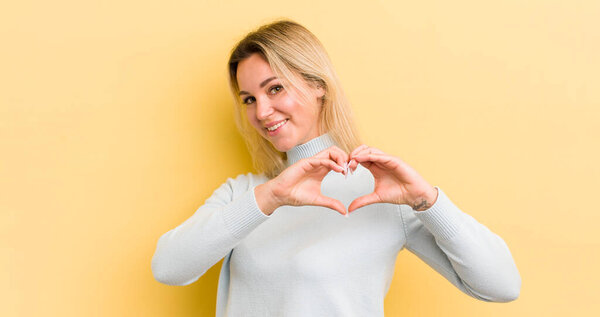 блондинка-кавказка улыбается и чувствует себя счастливой, милой, романтичной и влюбленной, придавая форму сердцу обеими руками
