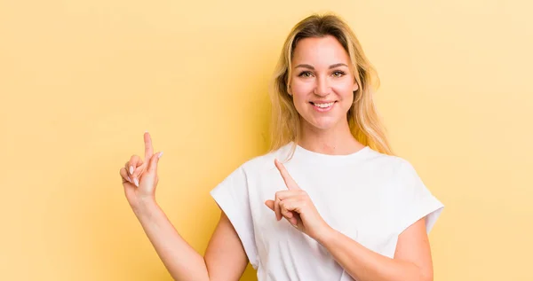 blonde caucasian woman smiling happily and pointing to side and upwards with both hands showing object in copy space