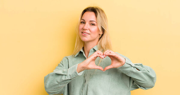 блондинка-кавказка улыбается и чувствует себя счастливой, милой, романтичной и влюбленной, придавая форму сердцу обеими руками