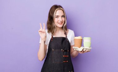 Güzel, beyaz bir kadın gülümsüyor ve arkadaş canlısı görünüyor. İki numarayı gösteriyor. kahve konseptini kaldır.