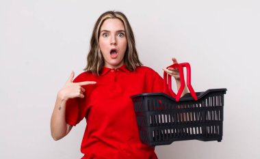 Ağzı açık, kendini işaret eden oldukça beyaz bir kadın şok olmuş ve şaşırmış görünüyor. boş alışveriş sepeti kavramı