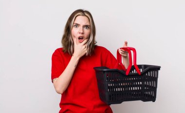 Ağzı ve gözleri açık, eli çenesinde güzel beyaz bir kadın. boş alışveriş sepeti kavramı