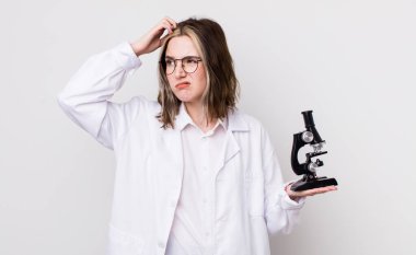 Kafası karışmış, kafası karışmış, kaşınan beyaz bir kadın. Mikroskop konseptine sahip bilim adamı