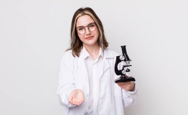 Güzel, beyaz bir kadın mutlu bir şekilde gülümsüyor ve bir konsept sunuyor. Mikroskop konseptine sahip bilim adamı