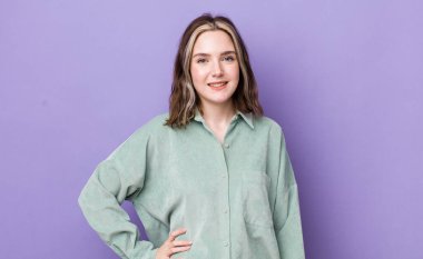 Güzel, beyaz bir kadın mutlu bir şekilde gülümsüyor, eli kalçasında ve kendinden emin, olumlu, gururlu ve arkadaş canlısı.