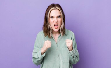 pretty caucasian woman shouting aggressively with annoyed, frustrated, angry look and tight fists, feeling furious