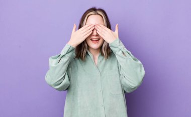 pretty caucasian woman smiling and feeling happy, covering eyes with both hands and waiting for unbelievable surprise