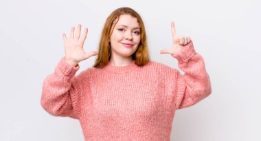 Güzel kızıl saçlı kadın gülümsüyor ve arkadaş canlısı görünüyor, 7 ya da 7 numarayı gösteriyor, eli önde, geri sayıyor.