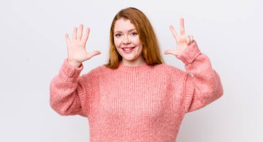 Güzel kızıl saçlı kadın gülümsüyor ve arkadaş canlısı görünüyor, 8 ya da 8 numarayı gösteriyor, eli önde, geri sayıyor.