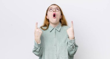 Güzel kızıl saçlı kadın şaşkın, şaşkın ve ağzı açık görünüyor, uzayı kopyalamak için iki elini de yukarı kaldırıyor.