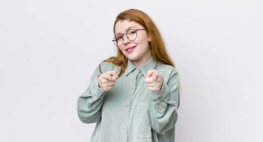 Güzel, kızıl saçlı, pozitif, başarılı, mutlu bir tavırla gülümseyen bir kadın kamerayı işaret ederek, elleriyle silah işareti yapıyor.