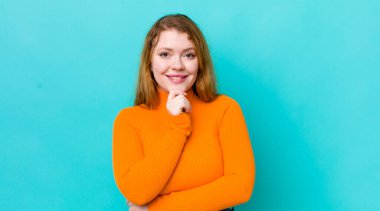 Güzel kızıl saçlı kadın mutlu ve gülümsüyor, elleri çenesinde, merak ediyor ya da soru soruyor, seçenekleri karşılaştırıyor.