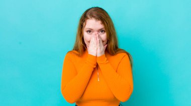 Güzel kızıl saçlı kadın mutlu, şanslı ve şaşkın görünüyor. İki eliyle ağzını kapatıyor.