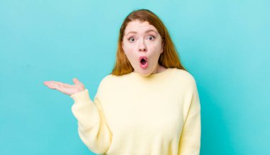 Güzel kızıl saçlı kadın şaşırmış ve şok olmuş görünüyor, çenesi düşmüş, eli açık bir objeyi tutuyor.