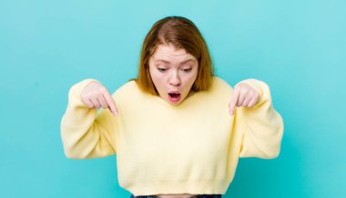 Güzel kızıl saçlı kadın şaşkın, ağzı açık ve şaşırmış hissediyor. İnançsızlık ve şaşkınlıkla aşağı bakıyor ve işaret ediyor.