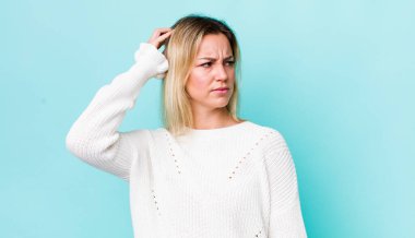 Güzel sarışın kadın şaşkın ve şaşkın hissediyor, başını kaşıyor ve yan tarafa bakıyor.