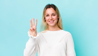 Güzel sarışın kadın gülümsüyor ve arkadaş canlısı görünüyor, üç ya da üç numarayı gösteriyor, eli önde, geriye doğru sayıyor.