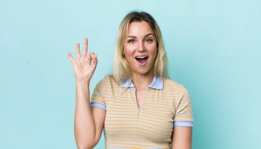 Güzel sarışın kadın kendini başarılı ve tatmin olmuş hissediyor, ağzı açık gülümsüyor, elle işaret veriyor.