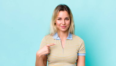 Güzel sarışın kadın gururlu, kendinden emin ve mutlu görünüyor, gülümsüyor ve kendini işaret ediyor veya bir numara işareti yapıyor.