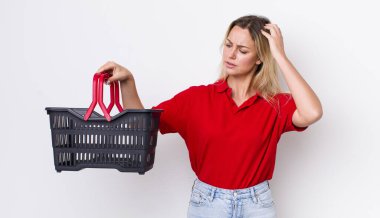 Kafası karışmış, kafası karışmış, kaşınan güzel sarışın bir kadın. Alışveriş sepeti konsepti