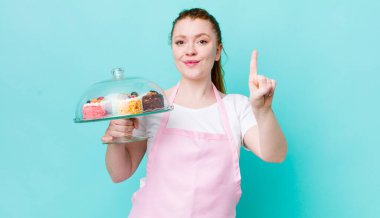 Kızıl saçlı güzel kadın gülümsüyor ve arkadaş canlısı görünüyor, bir numarayı gösteriyor. Ev yapımı kek konsepti