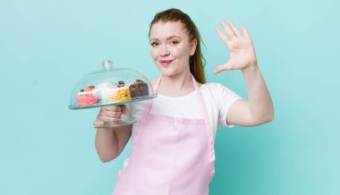 Kızıl saçlı güzel kadın gülümsüyor ve arkadaş canlısı görünüyor, beş numarayı gösteriyor. Ev yapımı kek konsepti