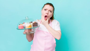 Kızıl saçlı, ağzı ve gözleri açık, eli çenesinde güzel bir kadın. Ev yapımı kek konsepti