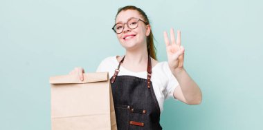 Kızıl saçlı güzel kadın gülümsüyor ve arkadaş canlısı görünüyor, üç numarayı gösteriyor. teslimat kavramını kaldır