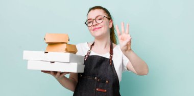 Kızıl saçlı güzel kadın gülümsüyor ve arkadaş canlısı görünüyor, üç numarayı gösteriyor. teslimat kavramını kaldır