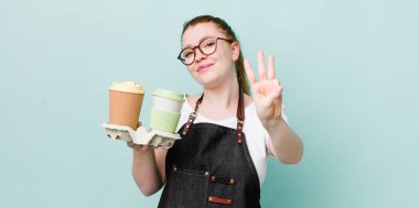 Kızıl saçlı güzel kadın gülümsüyor ve arkadaş canlısı görünüyor, üç numarayı gösteriyor. kahve konseptini kaldır.