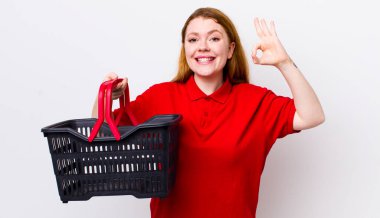 Kızıl saçlı güzel bir kadın kendini mutlu hissediyor. Onayını iyi bir hareketle gösteriyor. boş alışveriş sepeti kavramı