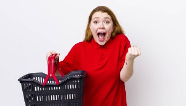 Kızıl saçlı güzel kadın şok olmuş hissediyor, gülüyor ve başarıyı kutluyor. boş alışveriş sepeti kavramı