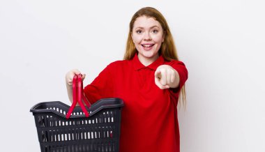 Kızıl saçlı güzel kadın kamerayı işaret ederek seni seçiyor. boş alışveriş sepeti kavramı