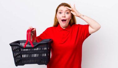 Kızıl saçlı güzel kadın mutlu, şaşkın ve şaşırmış görünüyor. boş alışveriş sepeti kavramı