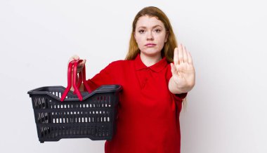 Kızıl saçlı güzel kadın ciddi görünüyor. Avuç içini gösteriyor. Durdurma hareketi yapıyor. boş alışveriş sepeti kavramı