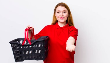 Kızıl saçlı, güzel bir kadın arkadaşça gülümsüyor ve bir konsept sunuyor. boş alışveriş sepeti kavramı