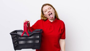 Kızıl saçlı, güzel, neşeli ve asi tavırlı, şaka yapan ve dilini dışarı çıkaran bir kadın. boş alışveriş sepeti kavramı