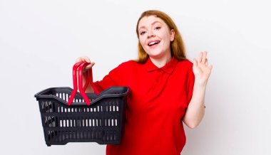 Kızıl saçlı güzel kadın mutlu bir şekilde gülümsüyor, el sallıyor, seni selamlıyor ve selamlıyor. boş alışveriş sepeti kavramı