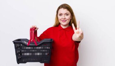 Kızıl saçlı güzel kadın gülümsüyor ve arkadaş canlısı görünüyor, iki numarayı gösteriyor. boş alışveriş sepeti kavramı